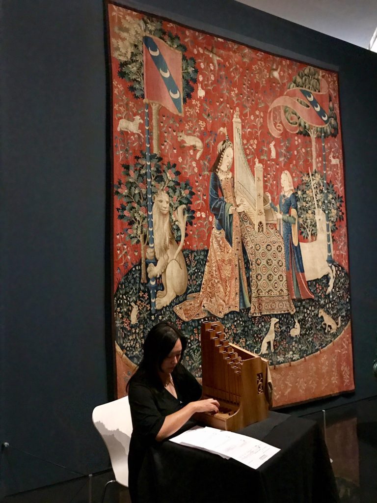 Photograph of musician Grace Chan playing the portative organ, alongside The lady and the unicorn tapestries at the Art Gallery of NSW.